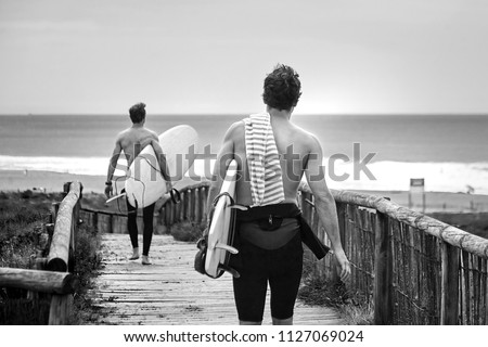 Two surfers running with surfboards on the beach. Men walking along the wooden path to the ocean to surf. Black and white image