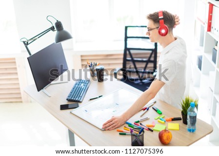 A young man with glasses and headphones stands near a computer desk and scratches his head. Before him lies a magnetic board and markers.