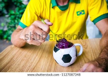 Soccer player in vintage brazil flag shirt sits eating acai from a football cup