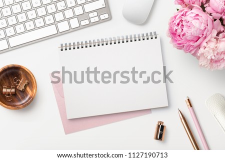 feminine workspace / desk with blank open notepad, keyboard, stylish office / writing supplies and pink peonies on a white background, top view Royalty-Free Stock Photo #1127190713