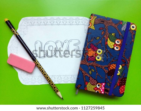 A sheet of paper with a painted word "love", a notebook with multi-colored owls, a pencil and an eraser lie on a yellow background.
