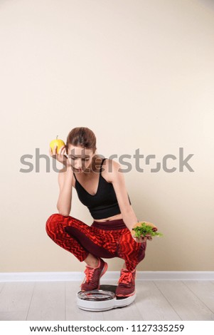 Woman holding tasty sandwich and apple while measuring her weight on floor scales near light wall. Weight loss motivation