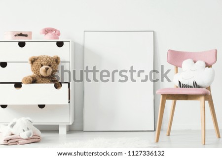 Empty poster with place for your picture standing on the floor in bright kid room interior with white cupboard and pink chair