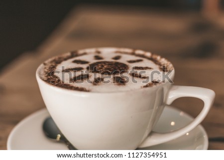 Cappuccino served in a white mug and saucer on a wooden table with chocolate sunshine decoration sprinkled in the frothy milk on top. Metal spoon on saucer, photo taken at 45 degrees.