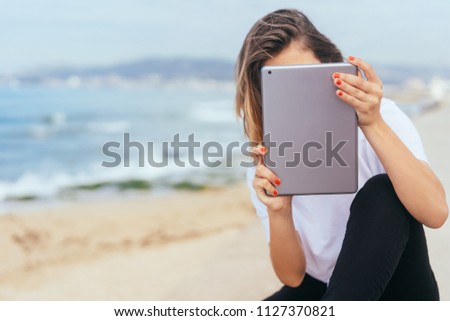 Young woman using a tablet at the seaside with her face hidden behind the screen and back to the ocean with copy space