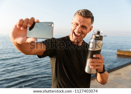 Image of handsome mature sportsman take a selfie with bottle of water on the beach.