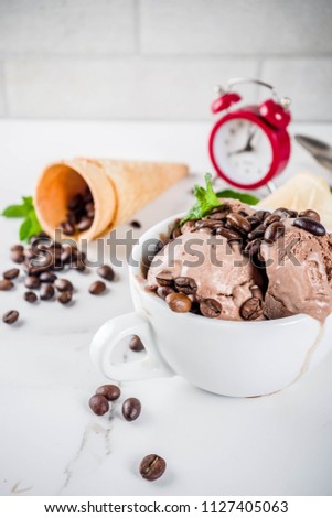Time for coffee concept, with an alarm clock for hours in the frame. Homemade coffee ice cream, served with coffee beans and mint leaves, with ice cream cones and spoons. White marble background,