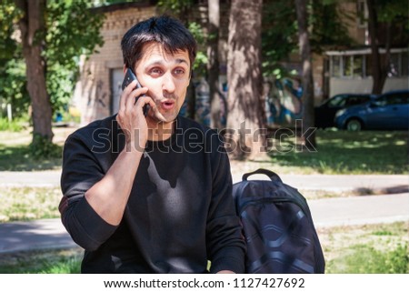 A young guy with a beard holds a smartphone and is emotionally surprised. A man is sitting on the bench with the phone