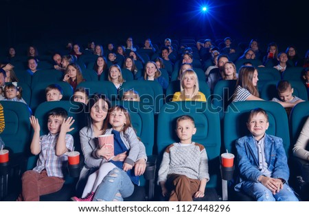 View of group of young people sitting in movie theatre, expressing emotions on their faces while watching film. Young spectators having fun, smiling. Project light source. 