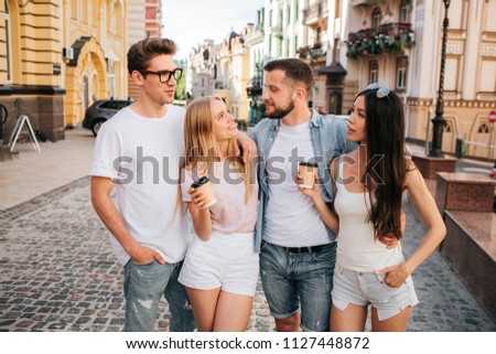 Interesting picture of girls holding cups of coffee and looking a bearded guy. He is stnading with his eyes closed. Guy in glasses is looking at chinese girl. They are standing together and posing.