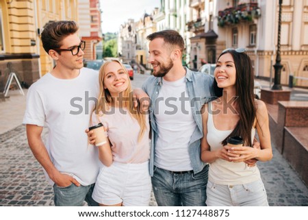 A picture of beautiful people standing together and posing. Blonde girl is smiling on camera and holding cup of coffee. Chinese girl is looking at guy in glasses. Bearded guy is looking at blonde girl