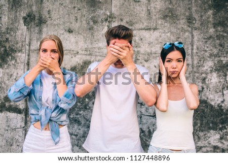 A picture of three young people standing together on grey background. Blonde girl is covering up her mouth with hands. Guy is covering up his eyes. Girl is covering up her ears with hands. They look