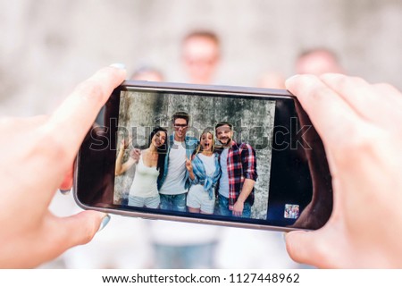 A picture of human's hands holding phone. This human is taking picture of four happy people. They are standing on grey background, posing and smiling.