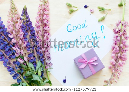 The inscription "Good morning" on a sheet of paper, lupine flowers and a gift on a wooden background.