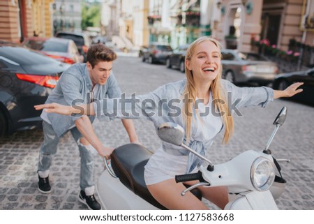 Excited and happy girl is holding her hands aside of body in air and keeping her eyes closed. Guy is standing behind her and trying to push motorcycle.