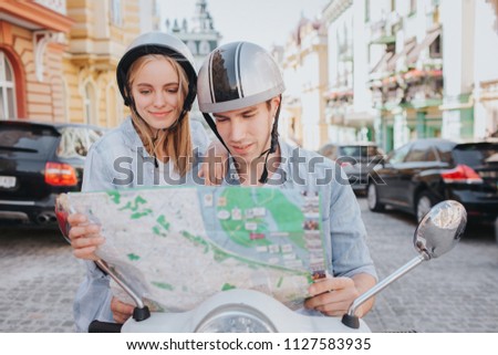 Concentrated and nice people are sitting on motorcycle and looking at map. Guy is holding it. They are standing in middle of road. They wear helmets.