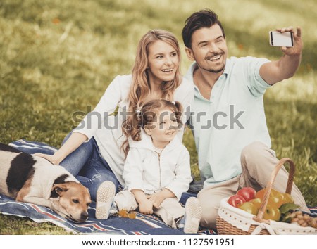 Father Takes Photo of Mother and Daughter on Smartphone on Picnic on Green Grass in Park on Sunny Summer Day. Happy Family and Healthy Lifestyle Concepts. Fun Outside in Summer Concept.