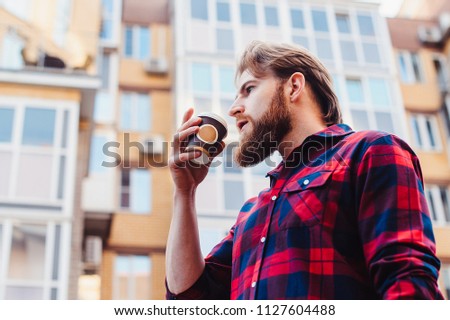 Lifestyle portrait concept. Bearded strolls on the streets of the city among the houses. Smoking and drinking coffee on the background of buildings