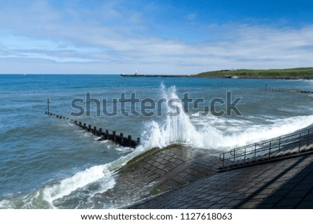 White foamy wave crashing on stone seawall on the coast of Aberdeen, Scotland, with jetty and lighthouses in the background
