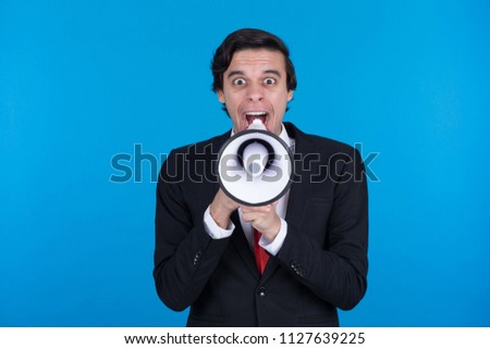 Close-up shot of crazy businessman wearing suit holding the megaphone on the front of his face shouting on blue background.