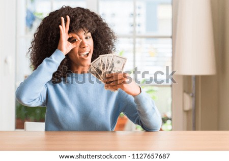 African american woman holding dollar bank notes with happy face smiling doing ok sign with hand on eye looking through fingers