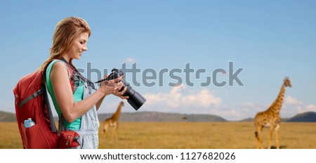 travel, tourism and photography concept - happy young woman with backpack and camera photographing over giraffe in african savannah background