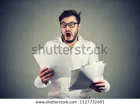 Young shocked guy in glasses looking at papers bank documents with list of bills and fees and feeling stressed on gray background Royalty-Free Stock Photo #1127732681