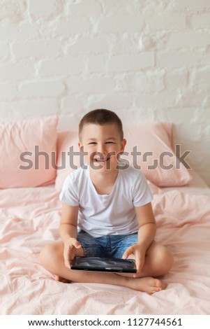Yong boy without a tooth in white shirt and blue jeans shorts sits on the bed in sunny light bedroom. Boy plays game tablet and smiles. Scrambled pink bed linen on the bed in Scandinavian interior 