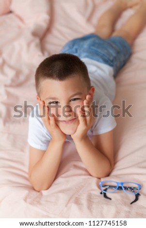 Young boy lies on the bed take off blue glasses wearing white shirt and jeans shorts props up his head. Pink bed linen in light sunny bedroom. 
