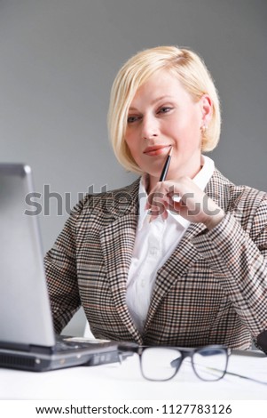 Beautiful blonde adult businesswoman dressed in checked stylish suit holding pen on table with laptop