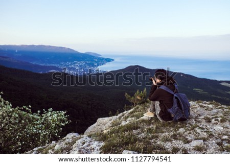 A traveler girl sits on top of a high mountain, takes pictures at the camera and looks at the sunset.