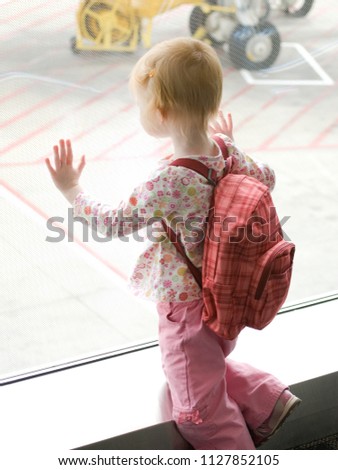 Little girl wearing pink pants and a red backpack presses hands on the airport window as she gazes at airplanes while waiting for her flight.