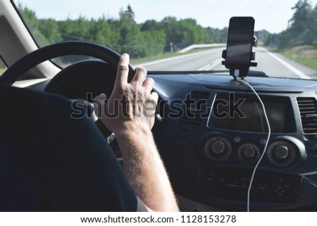 Man hand holding car steering wheel inside cabin with multimedia system, black dashboard and smartphone for navigation with road and green trees view from window.
