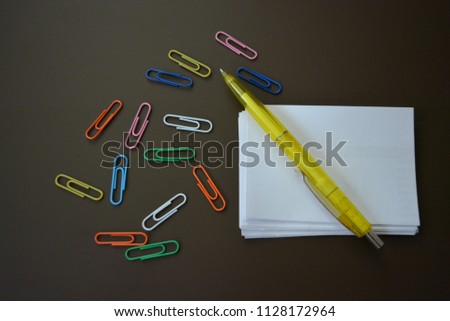 Color paperclips of different colors with sheets of white paper for recording and a yellow pen on a brown matte background