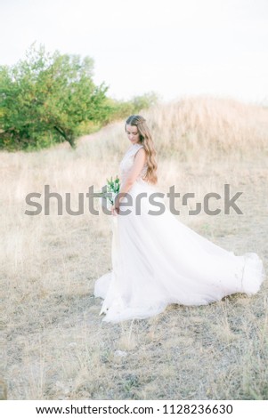 Beautiful young bride with long blond curly hair in a long white dress and bouquet in the summer field