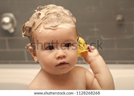 Children's hygiene. Small pretty baby boy sitting in bathroom with wet foam hair holding yellow duckling toy looking away, horizontal picture