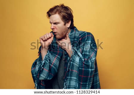 Young Sick Guy Wrapped in Checkered Plaid Coughing. Ill Handsome Person Feels Uncomfortably Unhealthy covering in Blue Wrap Isolated on Yellow Background. Concept of Cold Royalty-Free Stock Photo #1128329414