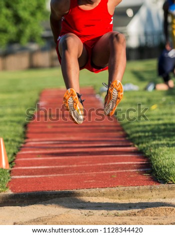 Long jumper in a red uniform is flying in the air ready himelf to land in the sand pit below. Royalty-Free Stock Photo #1128344420