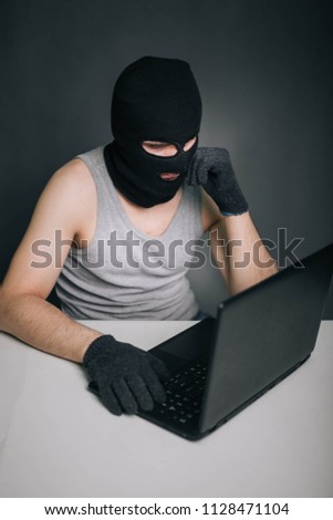 A thief in a black mask stole a laptop. A man in a balaclava and a computer in hand on a gray background. The hacker is hacking the computer. Steals information. A young man breaks the law.