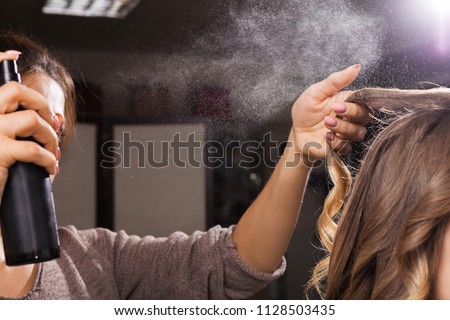hairdresser fixing a coiffure with ringlets of a young girl using a hair spray in a beauty salon. concept of professional stylist training Royalty-Free Stock Photo #1128503435