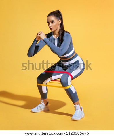 Sporty woman squatting doing sit-ups with resistance band. Photo of latin woman in fashionable sportswear on yellow background. Strength and motivation.