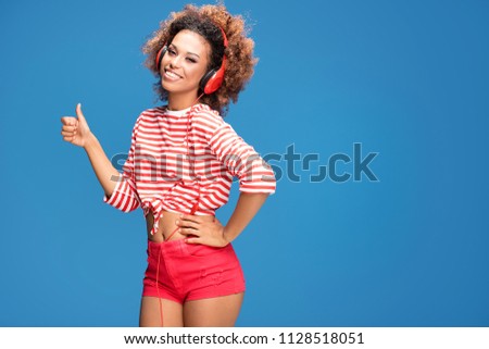 Adorable african american woman with red headphones smiling, looking at camera, blue background.