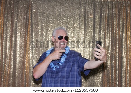 Selfies picture. A Man in a Photo Booth takes Selfie Photos with his cell phone as his photo is taken in a Photo Booth with a Gold Sequin Background. Photo Fun at Parties and Weddings.