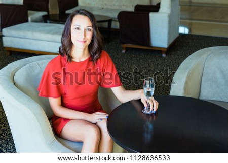 A girl is sitting in a hotel with a glass of water. The brunette in a red dress sits in a chair and holds a glass of water. The hotel lobby.
