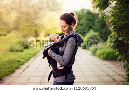 Little baby girl and her mother walking outside during sunset. Mother is holding and tickling her baby, babywearing in the ergo carrier Royalty-Free Stock Photo #1128695963