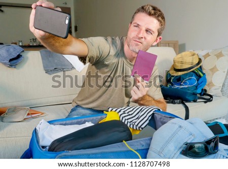 young happy and excited man taking selfie portrait with mobile phone camera showing passport while packing travel suitcase and preparing mess clothes  getting luggage ready for summer holidays 