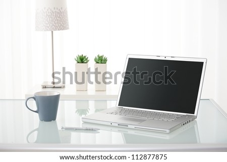 Still-life portrait of computer, pen, coffee mug on table, lamp and plants in bright environment. Royalty-Free Stock Photo #112877875
