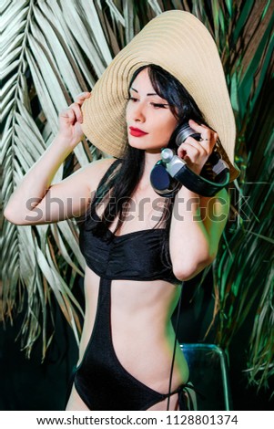 Brunette girl in bikini and thatched summer hat on the tropical leaves background. DJ woman plays music disko party on the equipment in the summer club.