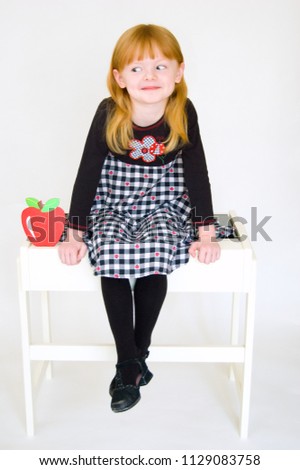 Cute young redheaded girl dressed in a black and white checked dress and black tights sits on top of a white school desk with a happy but sneaky expression on her face. Ready for back to school.