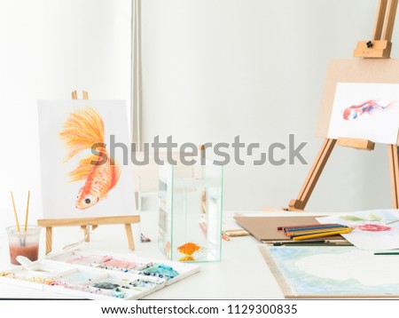 Wooden easel with a colorful pictures of goldfish and art supplies on table in the small art studio at home. 